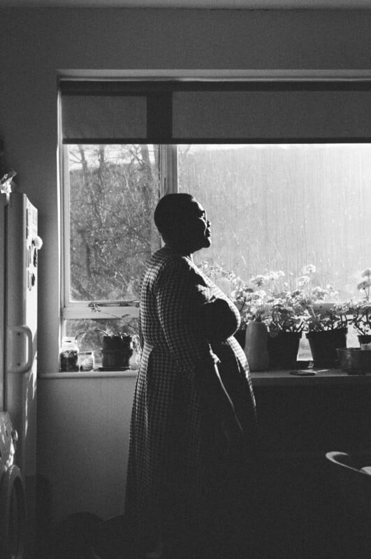 A person in a checkered dress stands in profile by a window filled with potted plants, bathed in soft natural light. The scene is in black and white, creating a calm and contemplative mood.