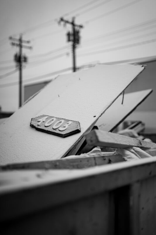 Una fotografía en blanco y negro que muestra un gran contenedor de basura lleno de escombros de construcción y una tabla de madera rota con el número 