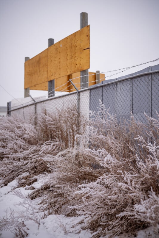 El heno cubierto de escarcha crece a lo largo de una cerca cubierta de alambre de púas; Detrás de la valla hay un cartel de madera sin terminar o desgastado por la intemperie, con nieve en el suelo bajo un cielo gris.