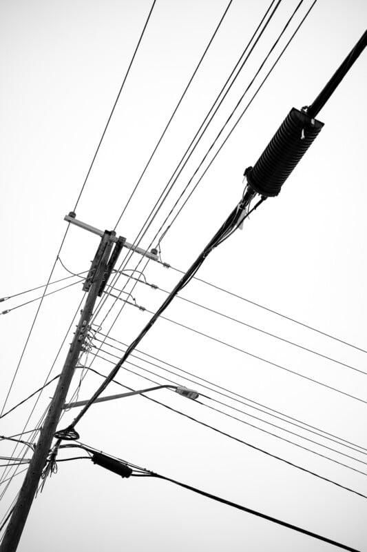 Fotografía en blanco y negro de postes y cables de servicios públicos que se cruzan en diagonal contra un cielo brillante y nublado. La imagen muestra varios cables, transformadores y farolas sujetos a postes.