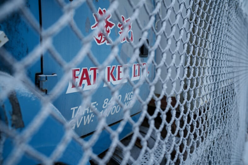 Primer plano de una valla de alambre de púas cubierta de nieve con una máquina industrial azul al fondo. El texto rojo y blanco y algunos números y letras son parcialmente visibles en la máquina.