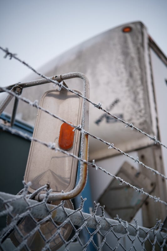 Primer plano de una valla de alambre esmerilado frente a un remolque viejo y desgastado con reflectores naranjas y luces cubiertas de nieve. La imagen está llena de una atmósfera fría e invernal.