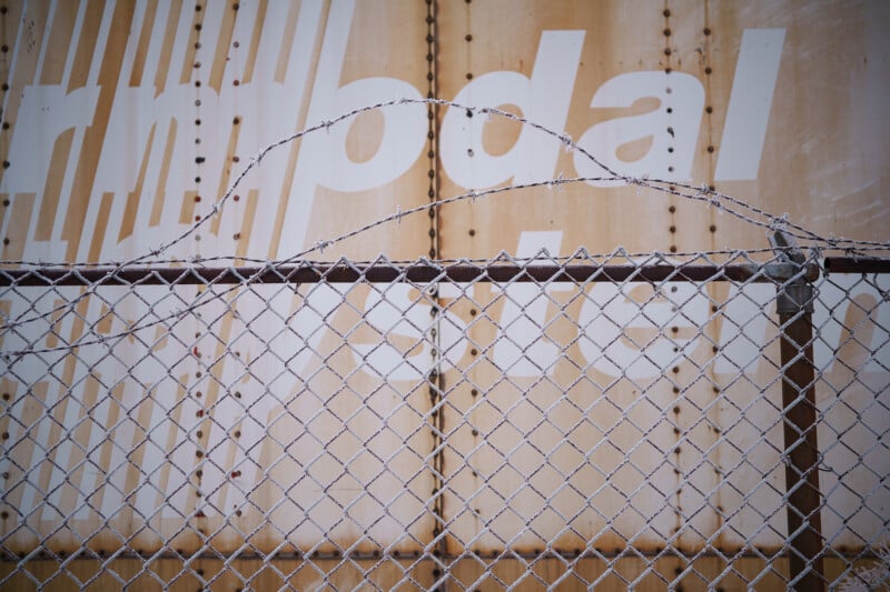 Frente a una pared de metal oxidado, pintada con grandes letras blancas que eran parcialmente visibles, había una valla de tela metálica rematada con una valla de tela metálica.