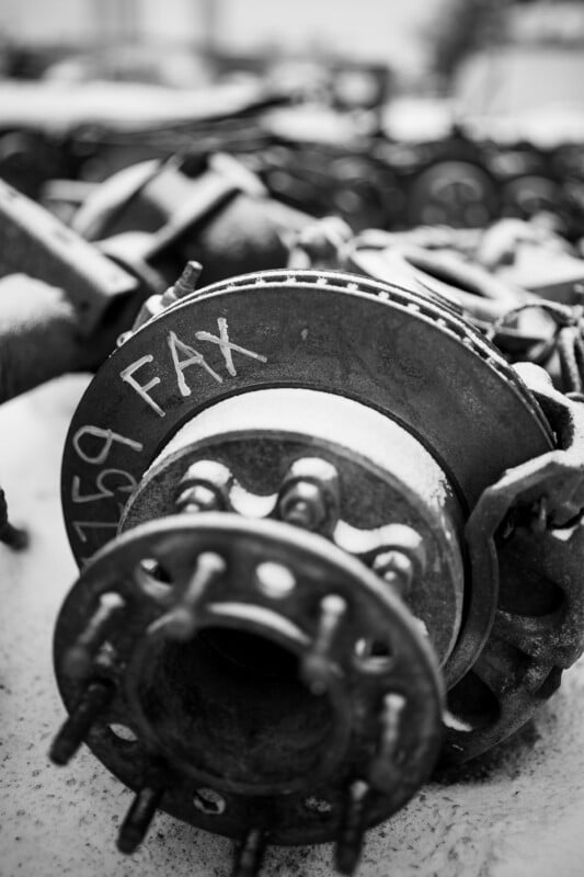 Fotografía en blanco y negro de un tambor de freno de vehículo oxidado con pernos y la palabra "fax" Escritura a mano en letras grandes sobre una superficie metálica, rodeada de otras piezas mecánicas.