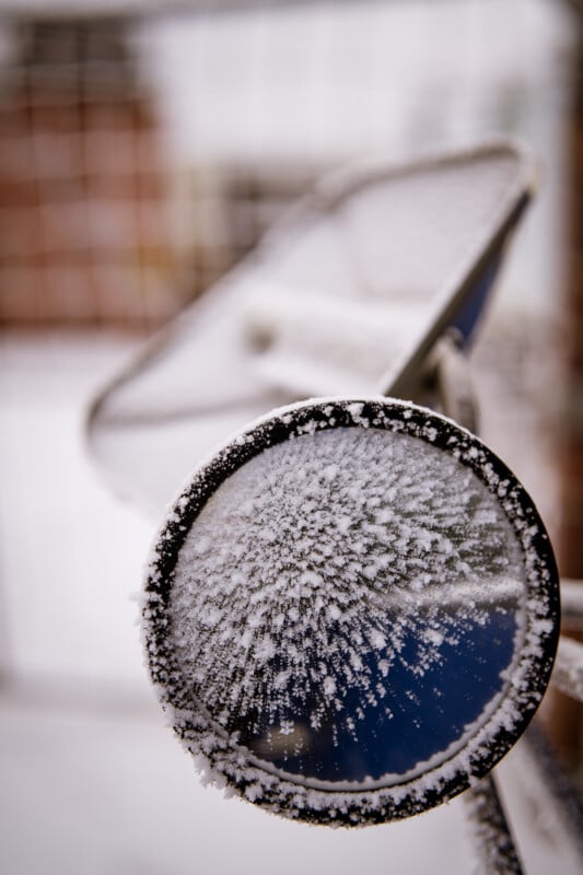 Primer plano de un espejo de bicicleta esmerilado cubierto de cristales de hielo, fondo de nieve borroso, valla metálica ligeramente desenfocada.