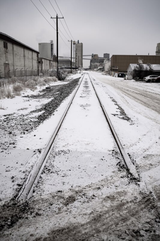 En un día nublado, las vías del ferrocarril cubiertas de nieve atraviesan una zona industrial llena de almacenes y postes telefónicos. Se podían ver neumáticos y huellas en la nieve junto a las huellas.