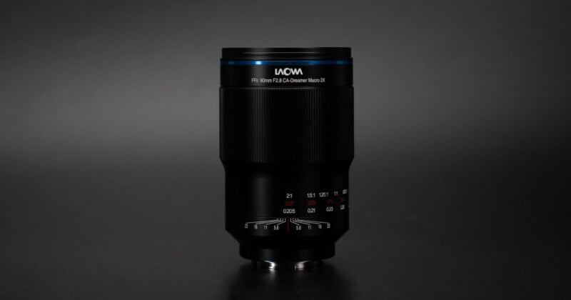 La lente de la cámara Laowa negra con detalles en azul se encuentra vertical sobre un fondo negro sólido, mostrando marcas de enfoque y apertura en el cilindro de la lente.