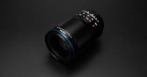 A black camera lens with blue accents and white and red markings sits on a dark surface, angled slightly, showing detailed text and measurement indicators on its barrel.
