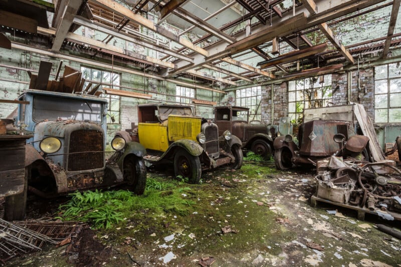 Four old, rusted vintage cars sit abandoned in a decaying building with broken windows and a mossy, debris-covered floor. Sunlight streams through gaps in the roof, highlighting overgrown plants inside.