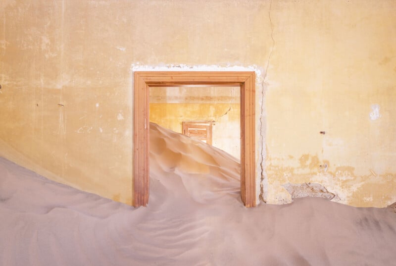 A series of doorways in an abandoned, yellow-painted building, partially filled with mounds of sand that flow through each room, creating a surreal, desert-like interior scene.
