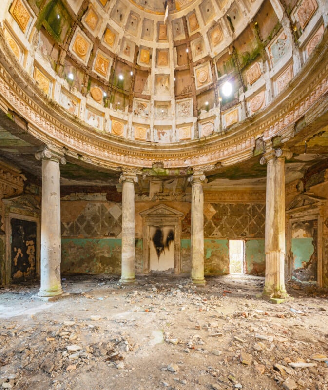 A wide view of a grand, abandoned domed hall with crumbling columns, peeling paint, debris on the floor, and sunlight streaming through a hole in the ornate ceiling. The space feels decayed yet architecturally impressive.