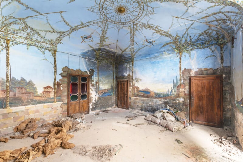 An abandoned room with peeling murals of outdoor scenes on the walls and ceiling, ornate wooden doors, crumbling plaster, debris, and sandbags scattered across the dusty floor.