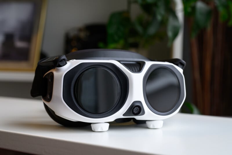 A pair of black and white virtual reality goggles with large round lenses sits on a white surface, with blurred plants and a framed photo in the background.