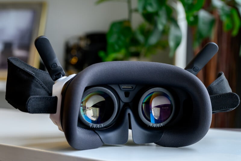 Close-up of a virtual reality headset resting on a white surface, with the lenses and interior padding visible. Reflections can be seen in the lenses, and a blurry green plant is in the background.
