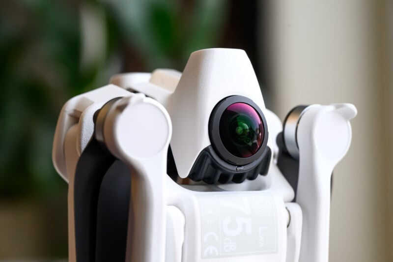 Close-up of a white drone camera with a wide lens, surrounded by mechanical arms, set against a blurred green background.