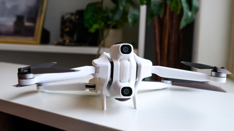A white quadcopter drone with black propellers sits on a white table. In the background, there are blurred objects, including a green plant and a framed picture.