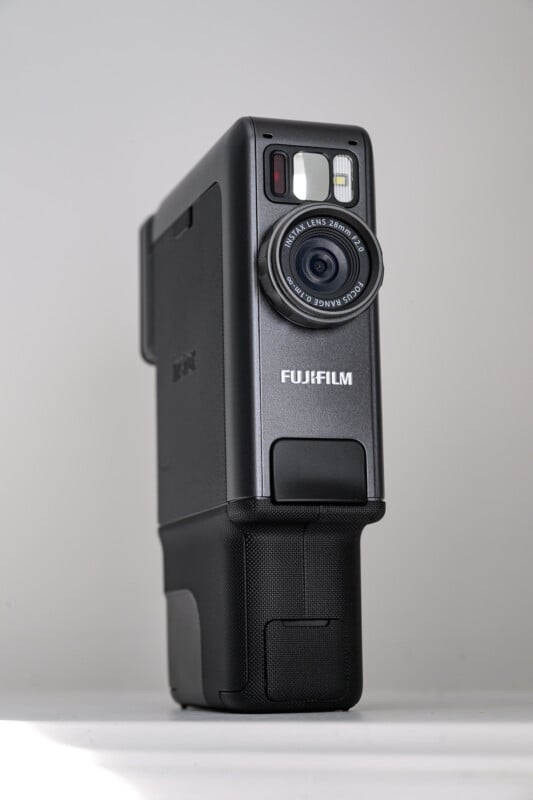 A Fujifilm instant camera is shown standing upright against a plain, light background. The camera lens and flash are visible, and the body is black with the Fujifilm logo prominently displayed.