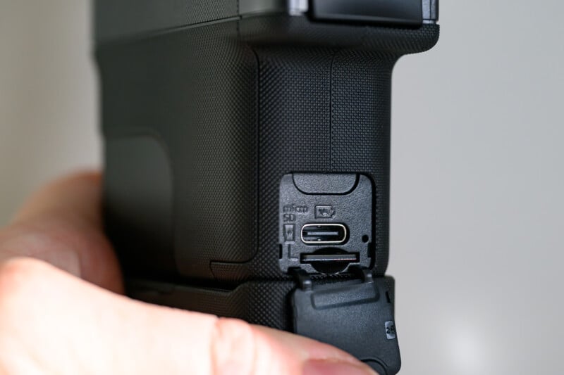 A close-up of a person's hand holding a camera, showing the open side port with slots for a microSD card and a USB-C connection.