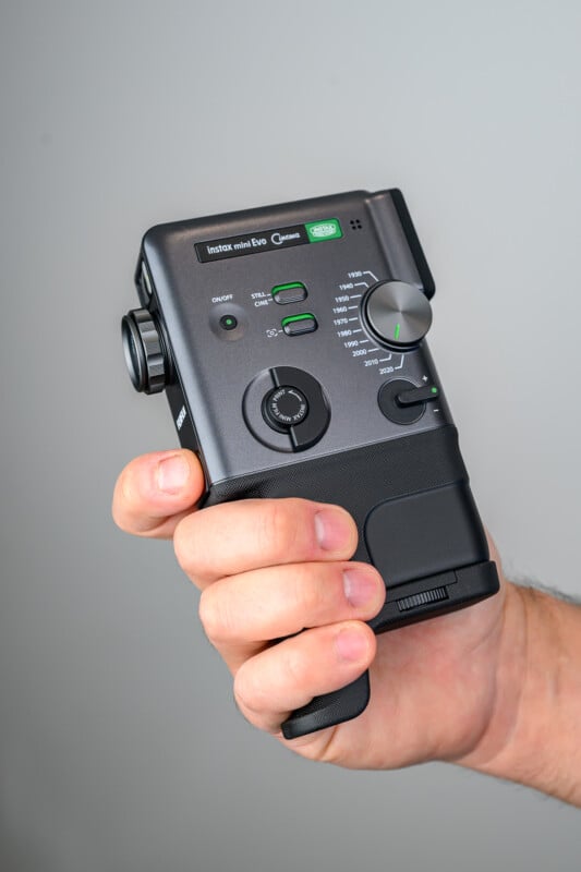 A hand holding a Fujifilm Instax Mini Evo instant camera against a plain gray background, showing the camera’s front buttons, dials, and lens.