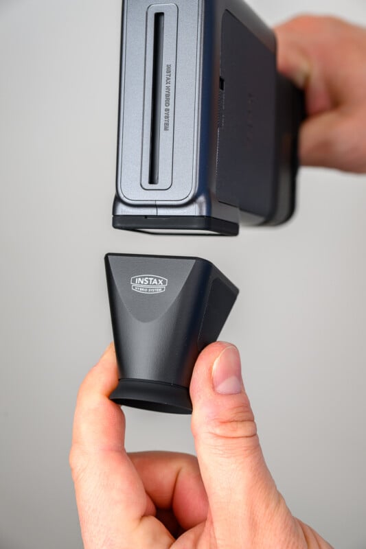 A close-up of hands holding and attaching an Instax camera accessory, aligning it beneath the slot of a gray Instax printer or camera device.