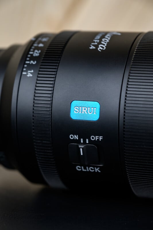 Primer plano de la lente de la cámara negra con azul "SIRUI" Logotipo, anillo de apertura e interruptores etiquetados. "Hacer clic" Tiene posiciones de encendido y apagado. El fondo está borroso.