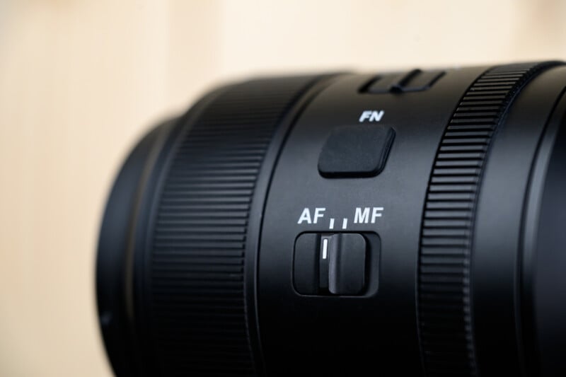 Primer plano de la lente de la cámara negra que muestra el interruptor etiquetado "Enfoque automático/enfoque manual" para enfoque automático y enfoque manual, y uno etiquetado "malla de fibra" Sobre fondo beige borroso.