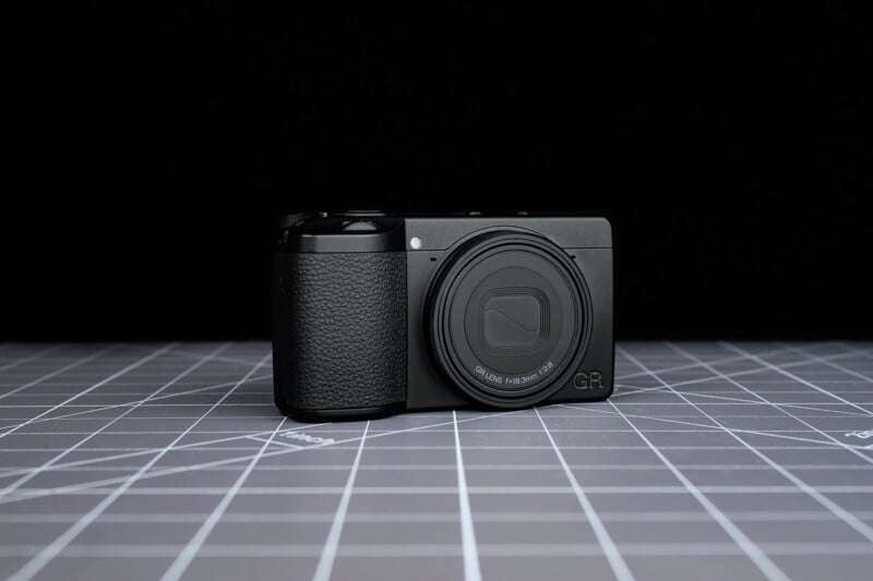 A compact black digital camera with a textured grip sits on a gray grid-patterned surface against a black background.
