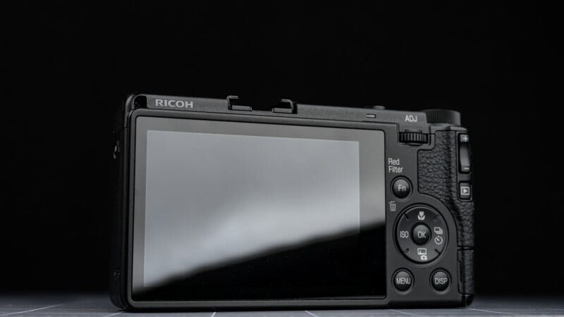 A close-up of the back of a Ricoh digital camera, showing its screen, control buttons, and textured grip against a plain black background.