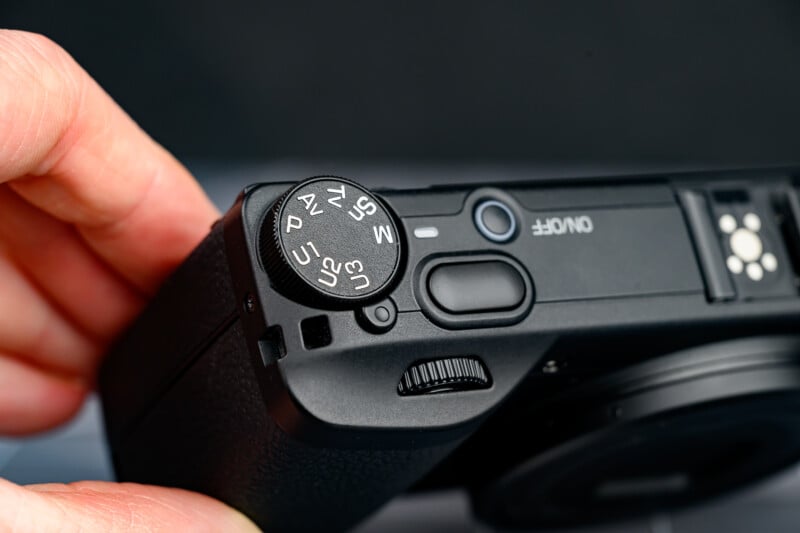 A close-up of a hand adjusting the mode dial on a black digital camera, showing settings like P, A, S, and M, with the on/off button and part of the camera lens visible.