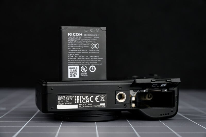 A Ricoh digital camera sits on a grid-patterned surface with its battery compartment open. The camera’s battery is partially inserted, standing upright above the compartment.