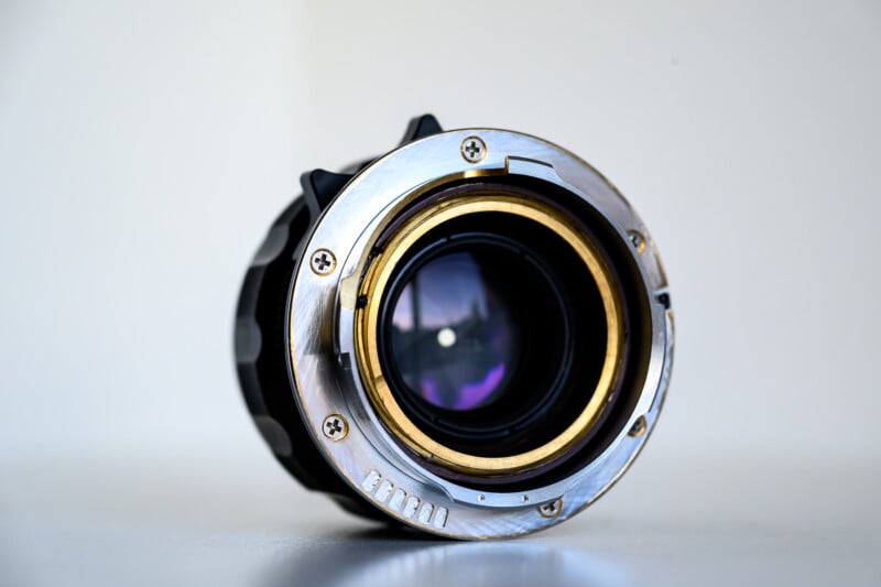 Close-up of the rear side of a camera lens with metallic mount, visible screws, and glass elements reflecting light, set against a plain white background.