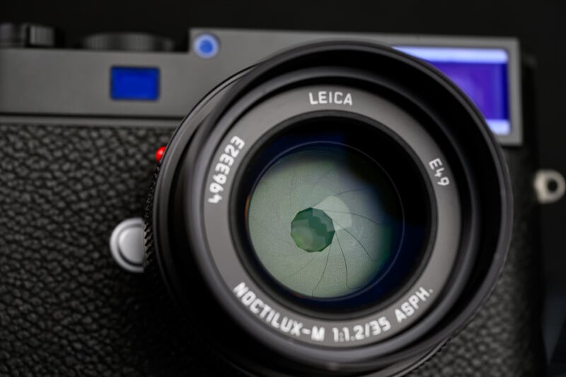 Close-up of a Leica camera lens showing the aperture blades. The lens markings read "NOCTILUX-M 1:1.2/35 ASPH." The camera body is partially visible with a textured finish and blue highlights.