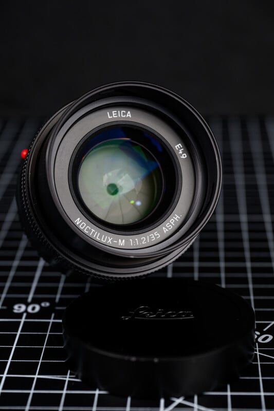 A Leica Noctilux-M 35mm f/1.2 ASPH camera lens is placed upright on a black grid-pattern surface, with its lens cap lying in front of it. The lens branding and details are clearly visible.