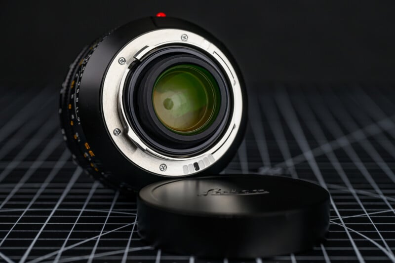 A camera lens facing upward rests on a grid-patterned surface, with its rear element and metal mount visible. The detached lens cap, labeled "Leica," lies nearby. The background is dark and out of focus.