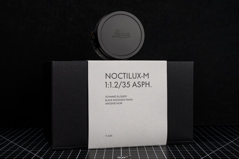 A black Leica camera lens with a cap stands on top of a black box labeled "NOCTILUX-M 1:1.2/35 ASPH." on a white band, against a dark, grid-patterned background.