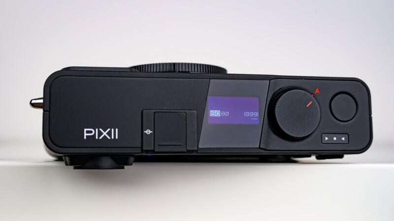 A close-up of the front of a black PIXII digital camera, featuring a small digital display, control dial, buttons, and a lens mount, all set against a plain white background.