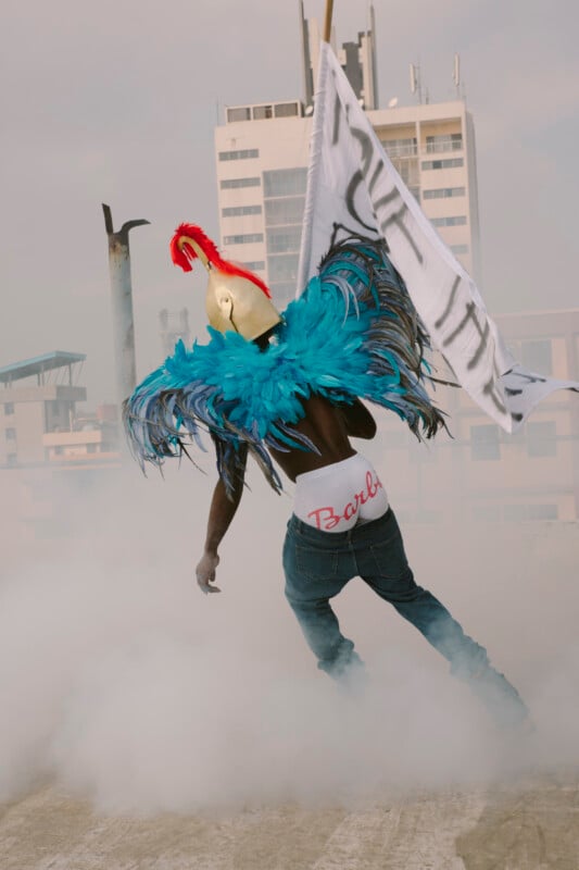 Un hombre que llevaba un casco dorado y alas de plumas azules. "Barbie" Un hombre en jeans y ropa interior está parado en una azotea rodeado de humo, sosteniendo una enorme bandera blanca con edificios al fondo.