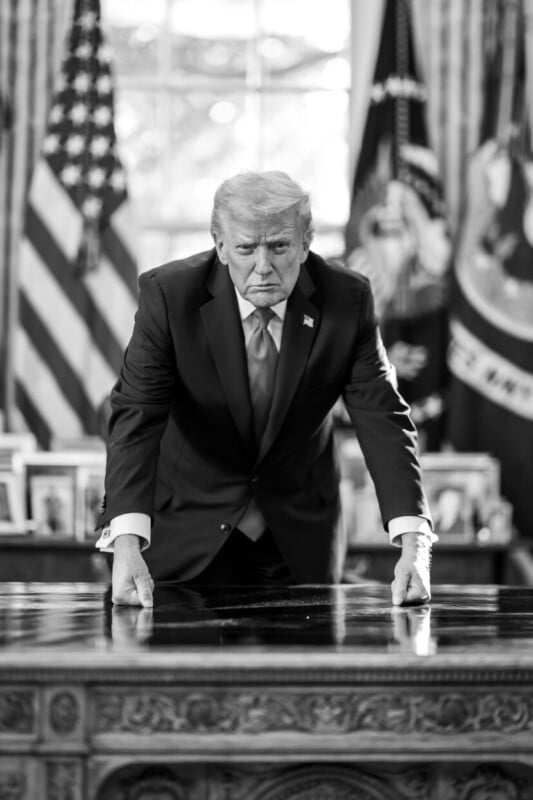 A man in a suit leans forward with both hands on a large desk, looking serious. Behind him are U.S. flags and framed photos, suggesting an official government office setting. The photo is in black and white.