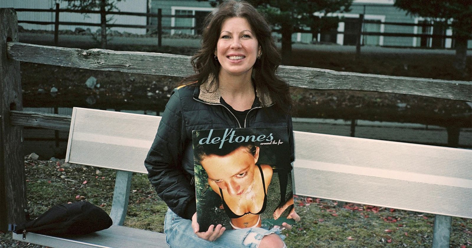 A smiling woman sits on a bench outdoors, holding a Deftones vinyl record in front of her. The album cover is positioned so it humorously aligns with her face and body. Trees and a fence are visible in the background.