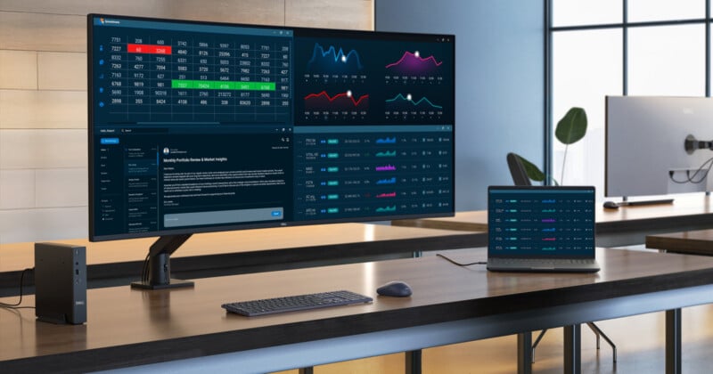A modern office desk with a large curved monitor displaying colorful charts, graphs, and data tables, next to a laptop showing similar data. A keyboard and mouse are on the desk. The background is a bright, minimal office space.
