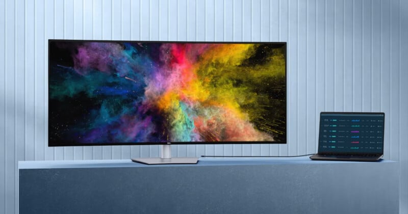 A widescreen monitor displaying a vibrant burst of colorful powder sits on a blue surface, connected by a cable to a slim black laptop showing graphs, against a light blue, vertical panel wall.