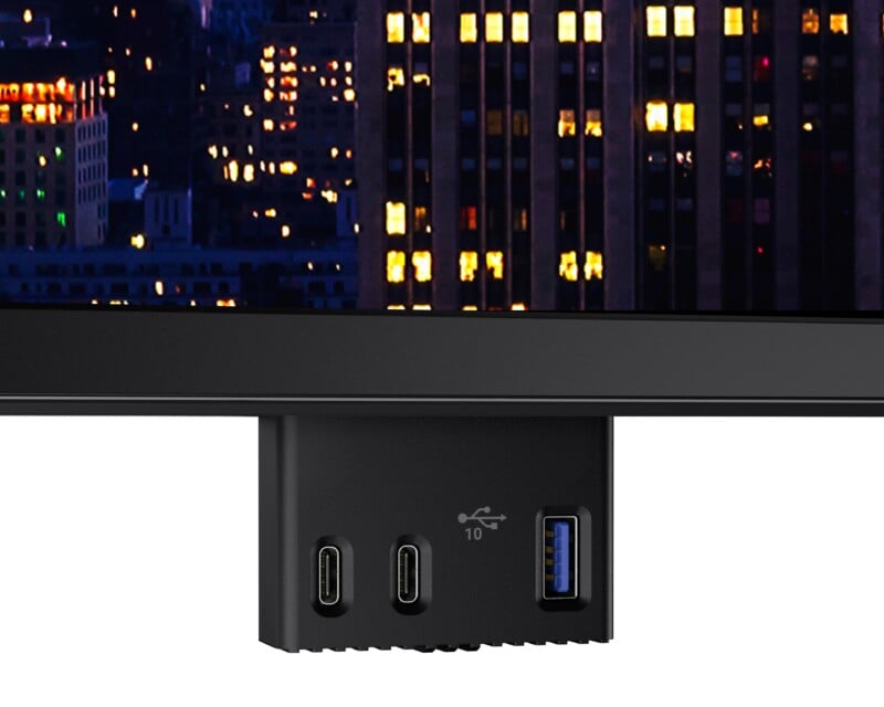 Close-up of the bottom edge of a monitor showing two USB-C ports and one blue USB-A port, with a cityscape illuminated by lights in the background on the screen.