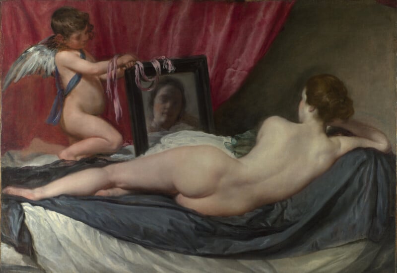 A nude woman reclines on a bed, facing away from the viewer, while a small winged child holds a mirror that reflects her face. The scene is set against draped pink and gray fabrics.