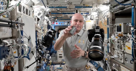 A person in a gray polo shirt floats inside a space station module, holding a small metallic object, with large cameras and equipment suspended in the air around them. Wires and devices cover the walls.