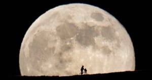 A person on a bicycle with a child silhouetted against a large, bright full moon in the night sky.