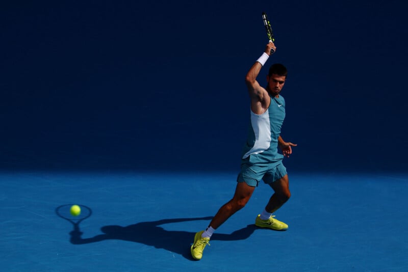 A tennis player in blue athletic wear and yellow shoes prepares to hit a forehand shot on a blue court, with his shadow and the ball visible on the ground.