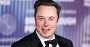 A man in a black tuxedo with a bow tie smiles at the camera while standing in front of a purple and blue event backdrop with white text.