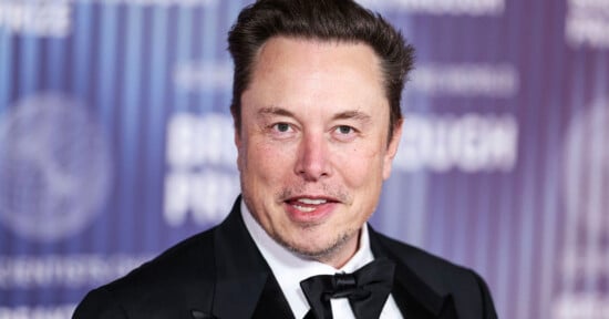 A man in a black tuxedo with a bow tie smiles at the camera while standing in front of a purple and blue event backdrop with white text.