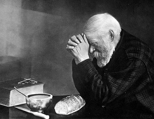 An elderly man with white hair and beard sits at a table, head bowed and hands clasped in prayer. In front of him are a loaf of bread, a bowl, a knife, and a large book with glasses resting on it.