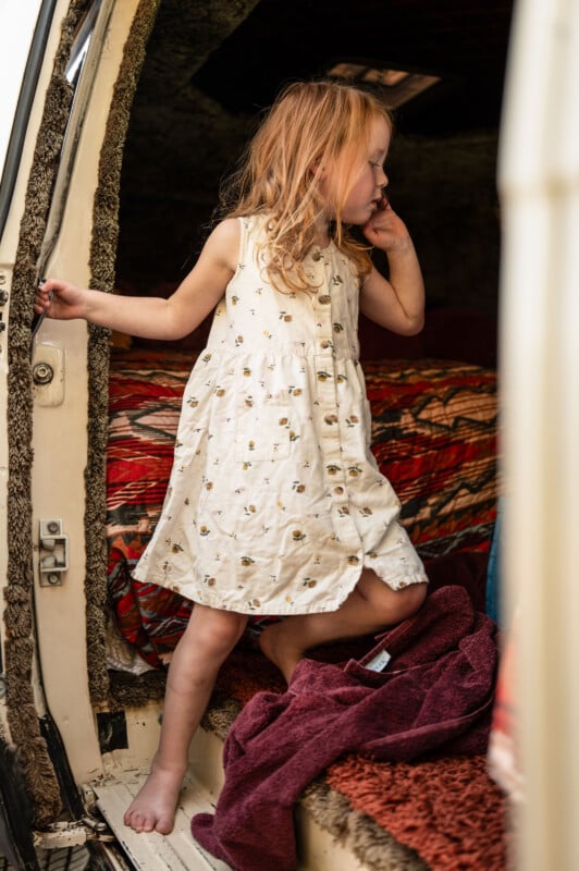 Una joven de largo cabello rubio, vestida con un vestido blanco con estampado floral, se paró en los escalones de la puerta abierta del auto y miró hacia un lado, rodeada de mantas y toallas de colores.